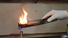 A gloved hand holds a block of ignited foam sealant over a bunson burner's flame.